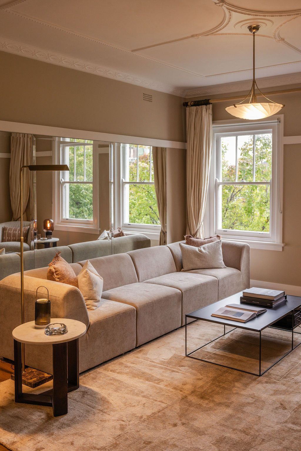 A warmly lit room with a large sofa, an antique lamp hanging off an adorned cieling in a relaxed setting with greenery-filled windows