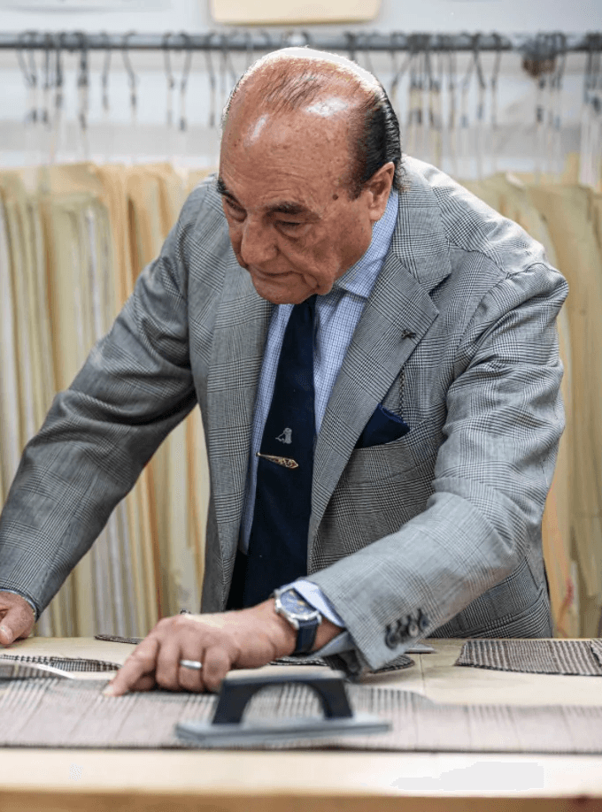 A tailor in a classic italian suit at his workbench pointing as another tailor cuts away at the garment, there is a garment rack in the background