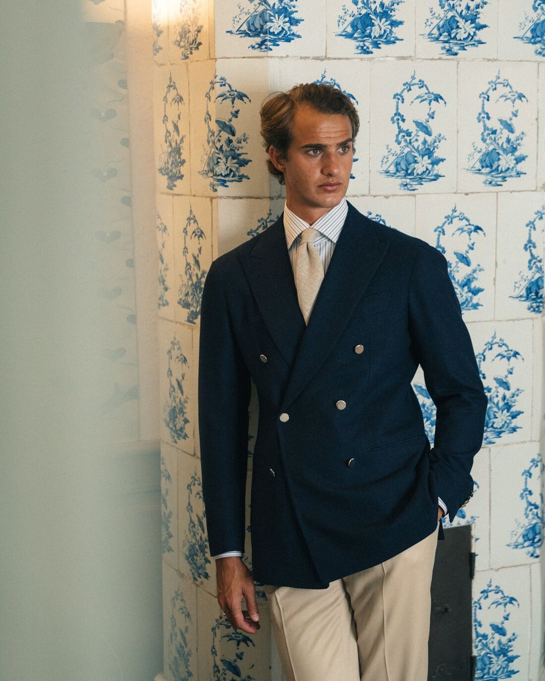 A man wearing a classic italian double-breasted suit resting his back on a wall made up of vintage floral-patterned bathroom tiles