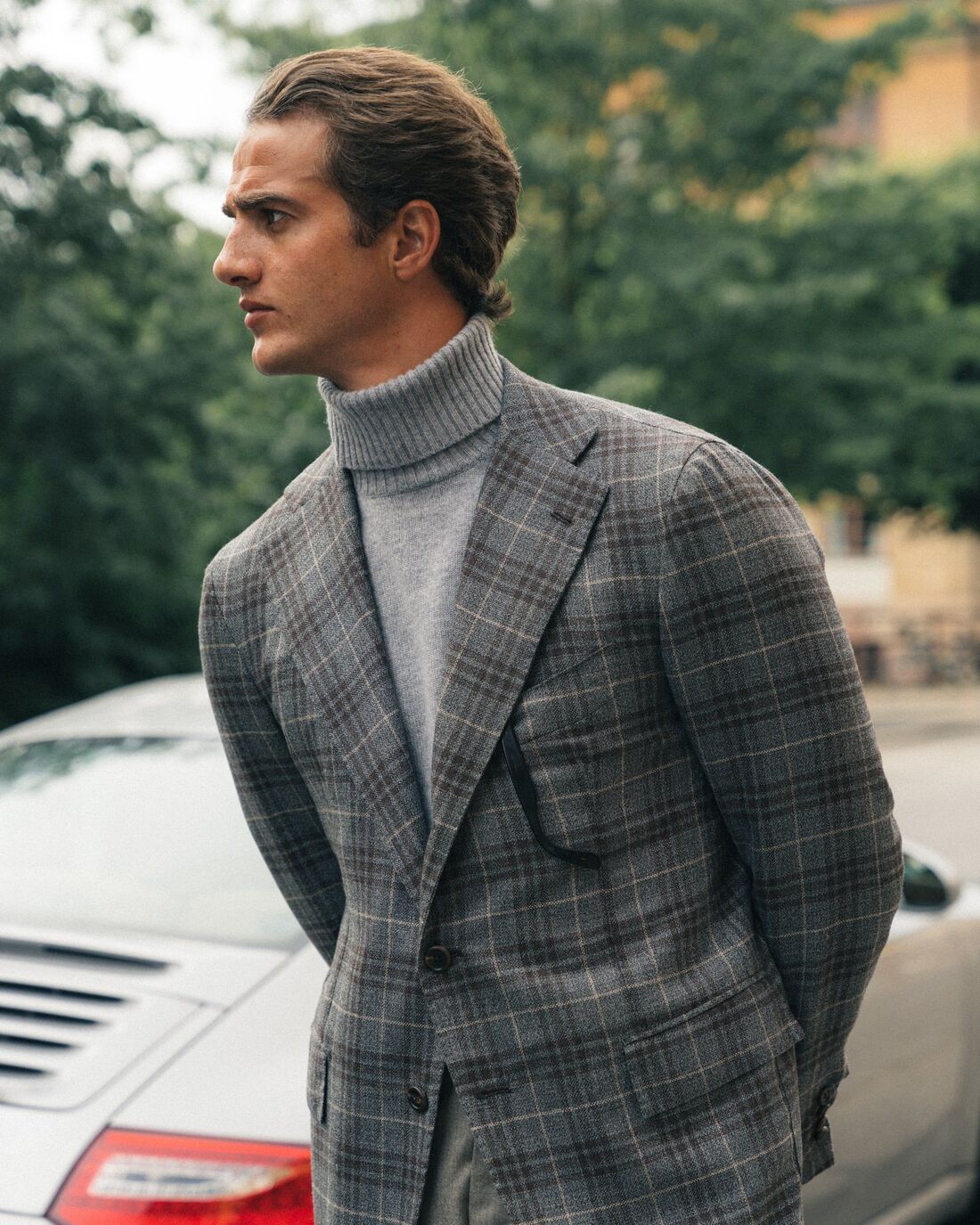 Mid-wait close-up of a man's profile, wearing a turtleneck and a checkered pattern suit, there is a Porsche coupe in the background & greenery