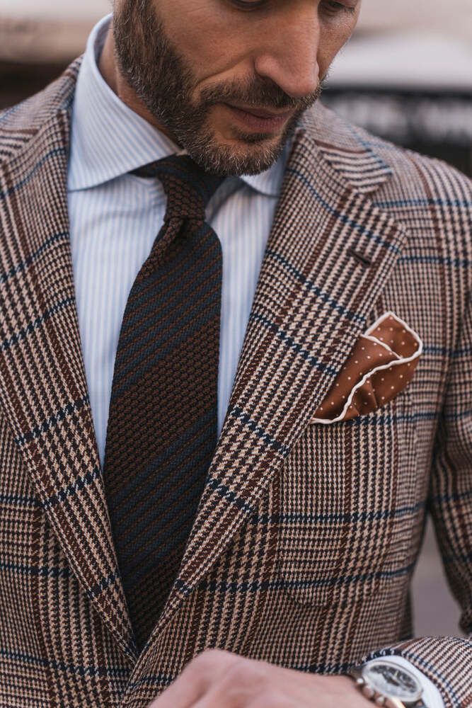 Close-up of a man wearing a checkered suit with a patterned tie, pinstripe shirt and a dotted handkerchief is looking at his watch