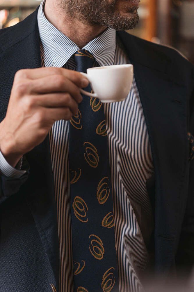 close-up of a suited man drinking an espresso, he's wearing a pinstripe shirt and a patterned tie