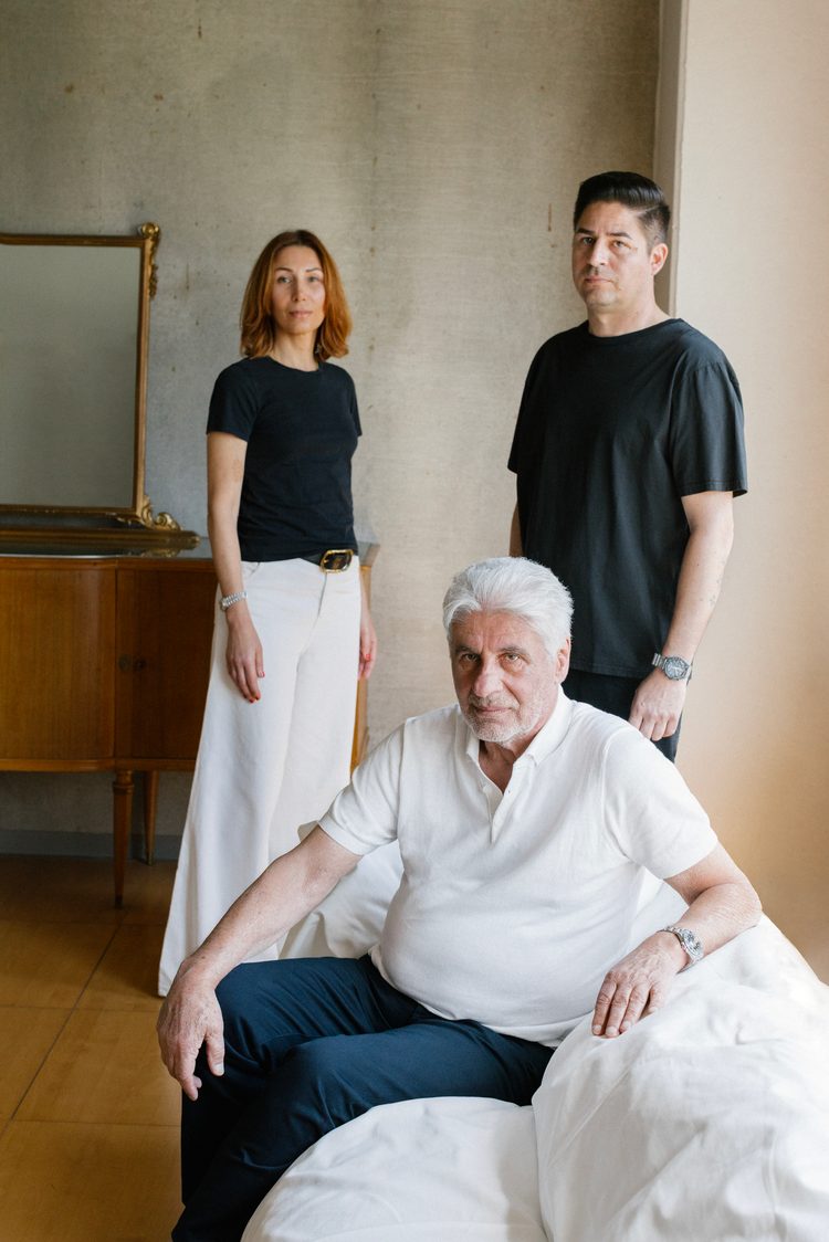 An older man in a white polo shirt seated on a white sofa, with a woman and a man in black standing behind him beside a gilded mirror