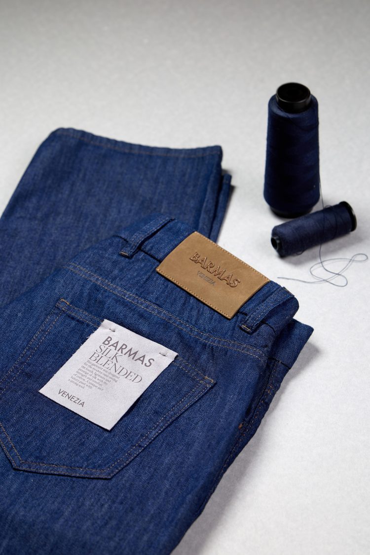A folded pair of indigo Barmas Venezia jeans with a tan leather patch and paper pocket label, beside two cones of navy thread
