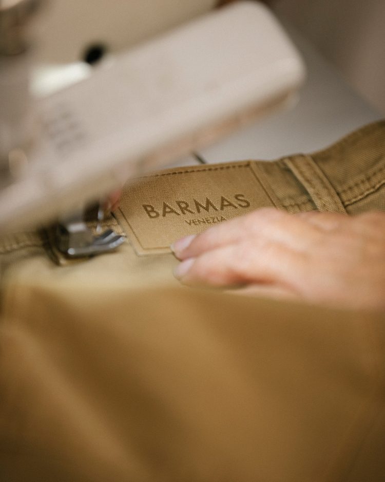 A close-up of hands at a sewing machine, guiding a Barmas Venezia woven label onto the waistband of tan denim
