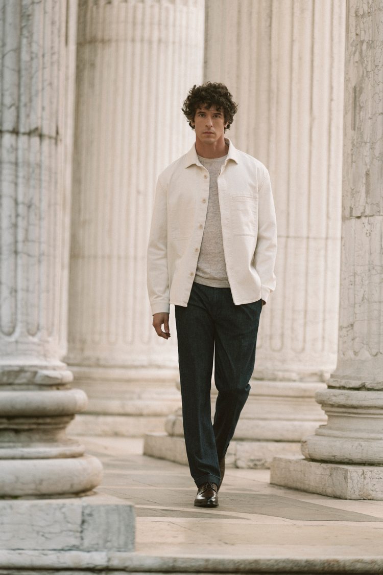 A man walking between towering marble columns in a cream overshirt and knit with dark denim trousers