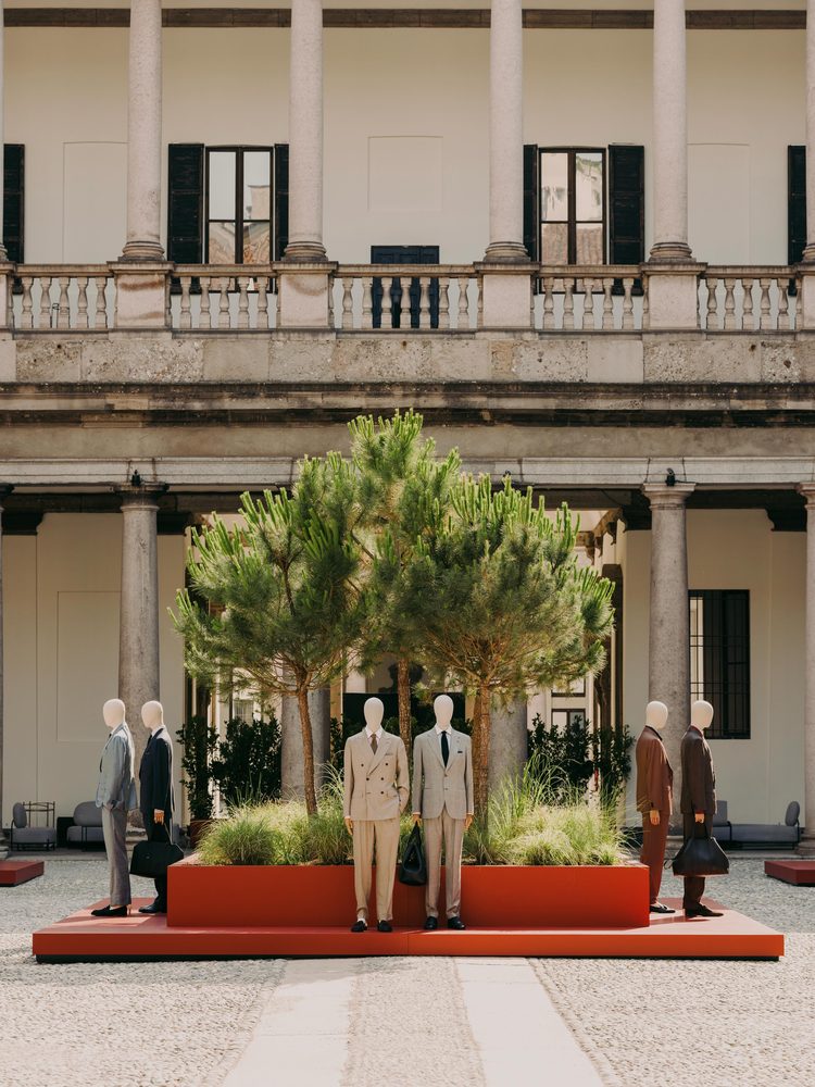 Mannequins dressed in tailored suits standing on low red platforms among pine trees, in the colonnaded courtyard of a historic palazzo