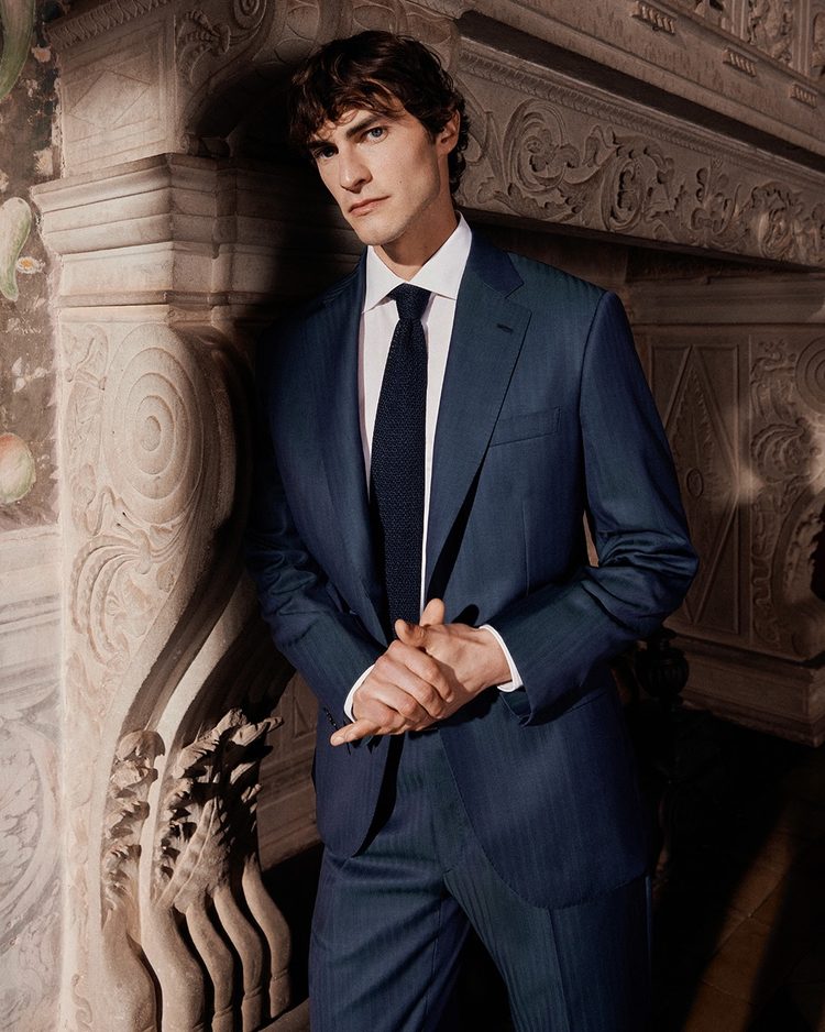 A man in a deep blue herringbone suit, white shirt and dark knitted tie, leaning against an ornately carved stone mantelpiece