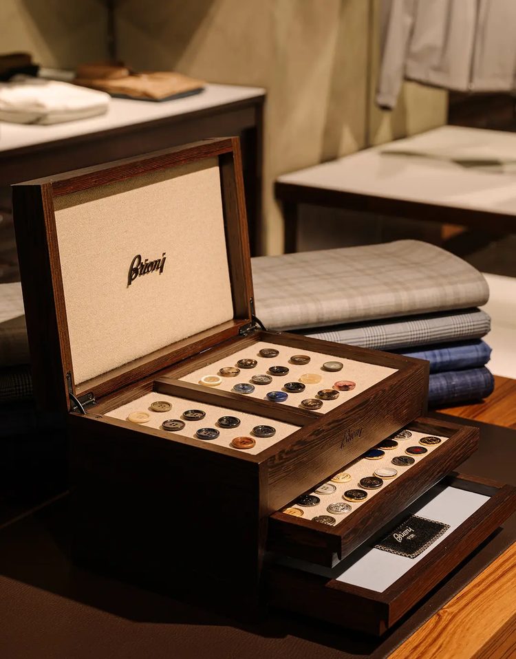 An open Brioni button chest in dark wood, its drawers holding rows of horn and mother-of-pearl buttons, with folded suiting fabrics stacked behind