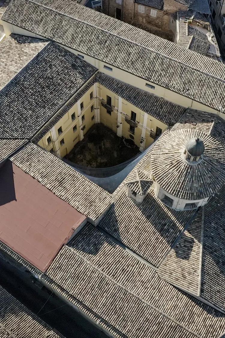 An aerial view of a historic Italian building with terracotta-tiled roofs, a small dome and an inner courtyard, in warm afternoon light