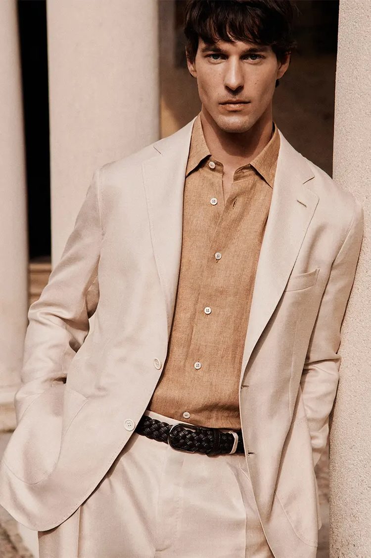 A man in a cream suit and tan linen shirt with a woven leather belt, leaning against a stone column
