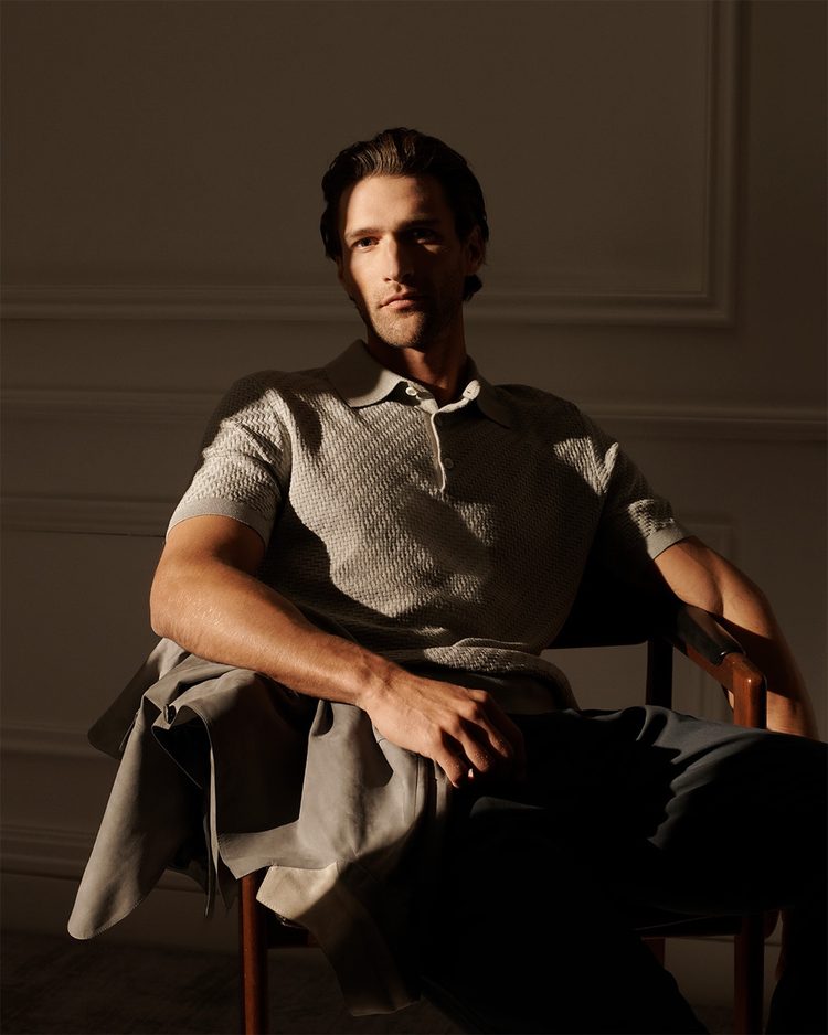 A man seated in a wooden armchair in a dim panelled room, wearing a textured knitted polo shirt, with a suede jacket draped across his lap