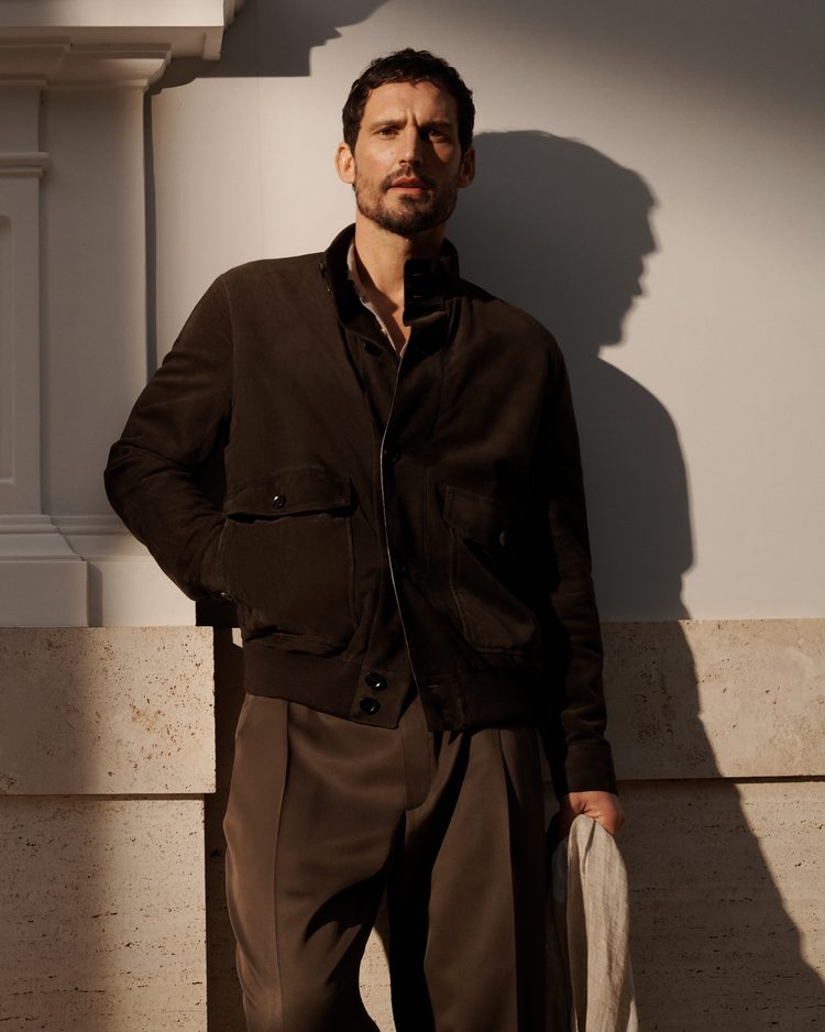 A man in a dark brown suede blouson with flap pockets and tobacco trousers, leaning against a sunlit white wall with a pale linen garment in hand