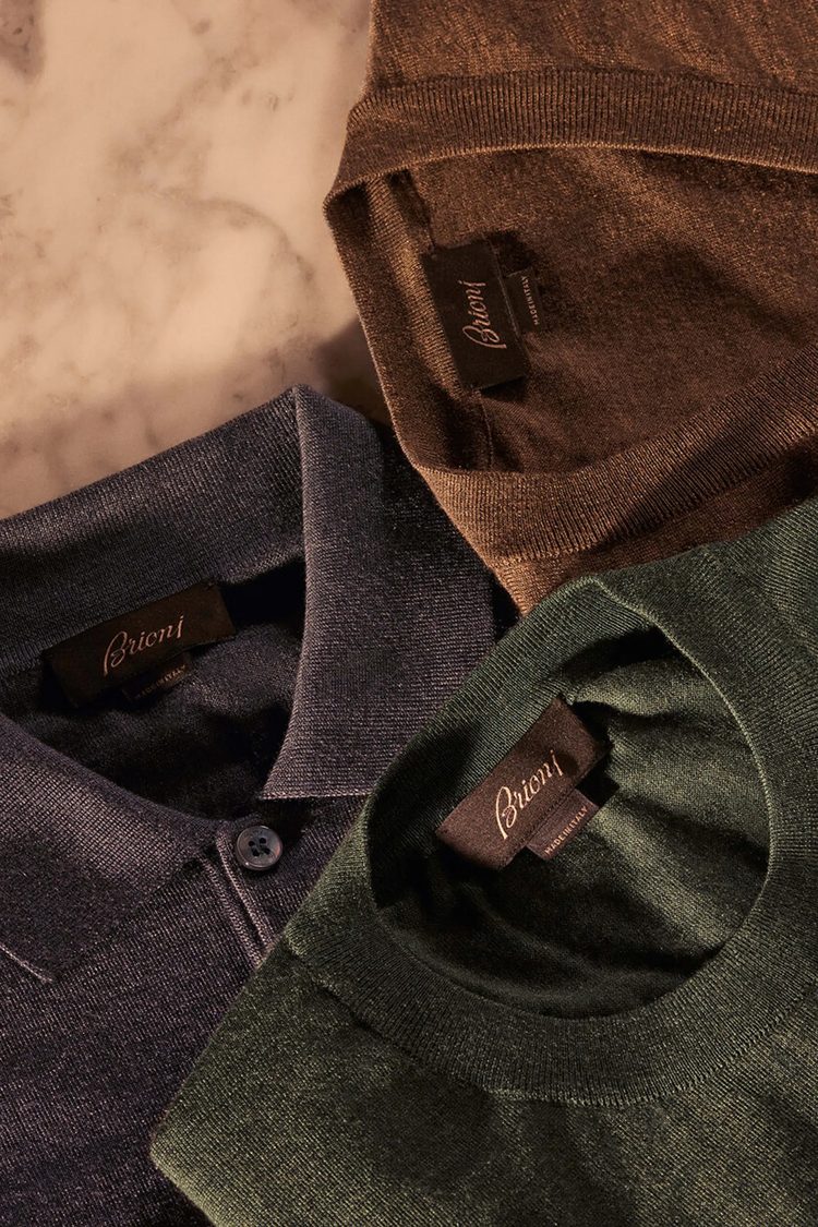 Three folded pieces of fine Brioni knitwear in brown, slate blue and forest green, layered on a marble surface