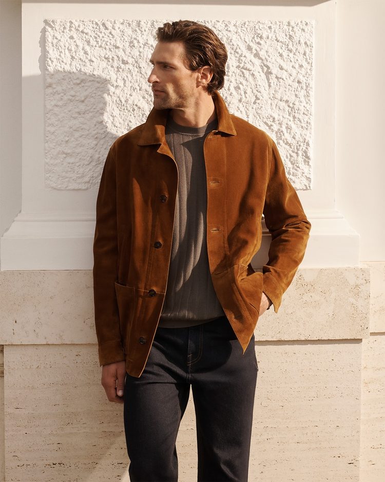 A man standing before a white stone wall in a tobacco suede jacket over a grey knitted crew neck and dark denim
