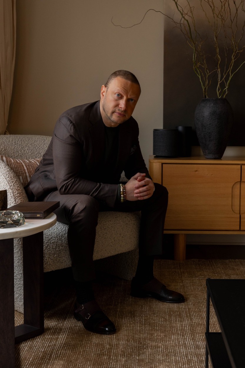 Deorum's founder seated in the Toorak showroom, in a dark brown suit with hands clasped, beside a wooden sideboard and dried branches in soft, warm light