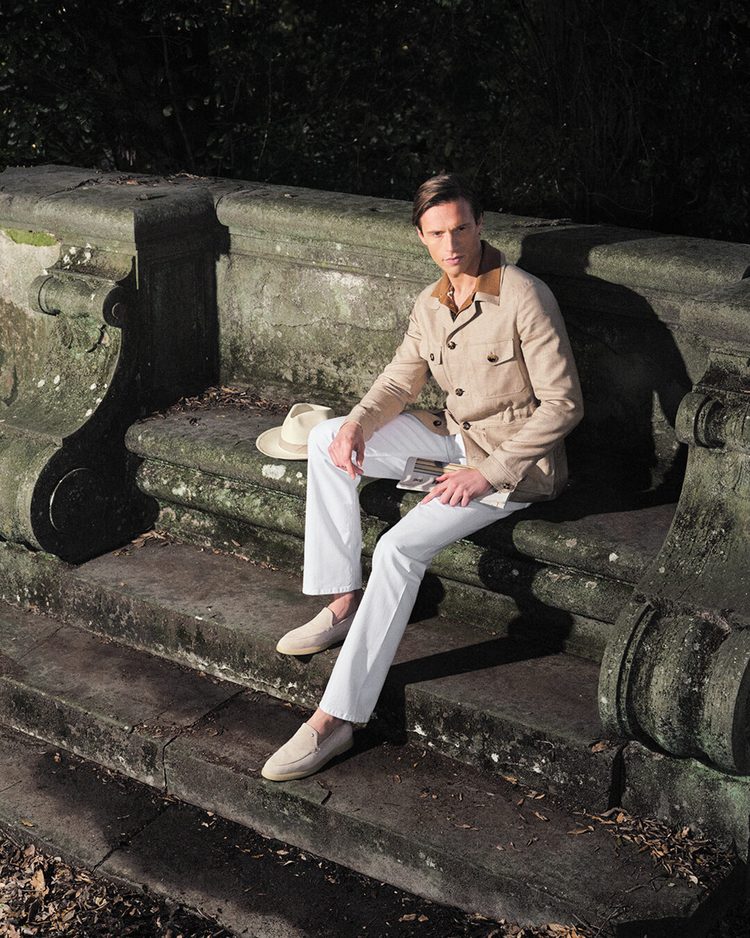 A man seated on a weathered stone garden bench in a beige linen safari jacket and white trousers, a panama hat resting beside him