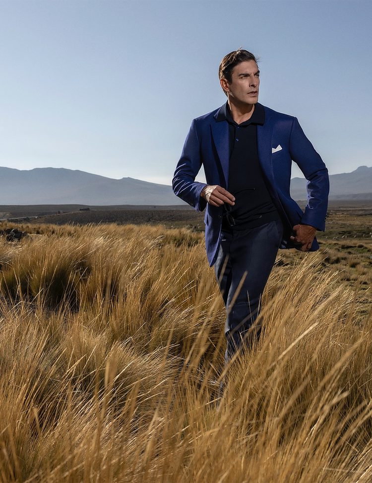 A man walking through tall golden grass beneath distant mountains, wearing a vivid blue jacket over a navy polo shirt with a white pocket square