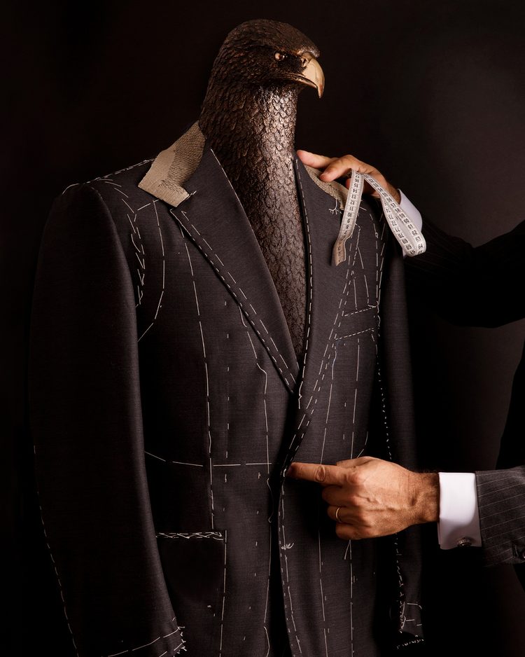 A tailor with a measuring tape fitting a basted suit jacket, marked with white tacking stitches, on a bronze eagle-headed mannequin