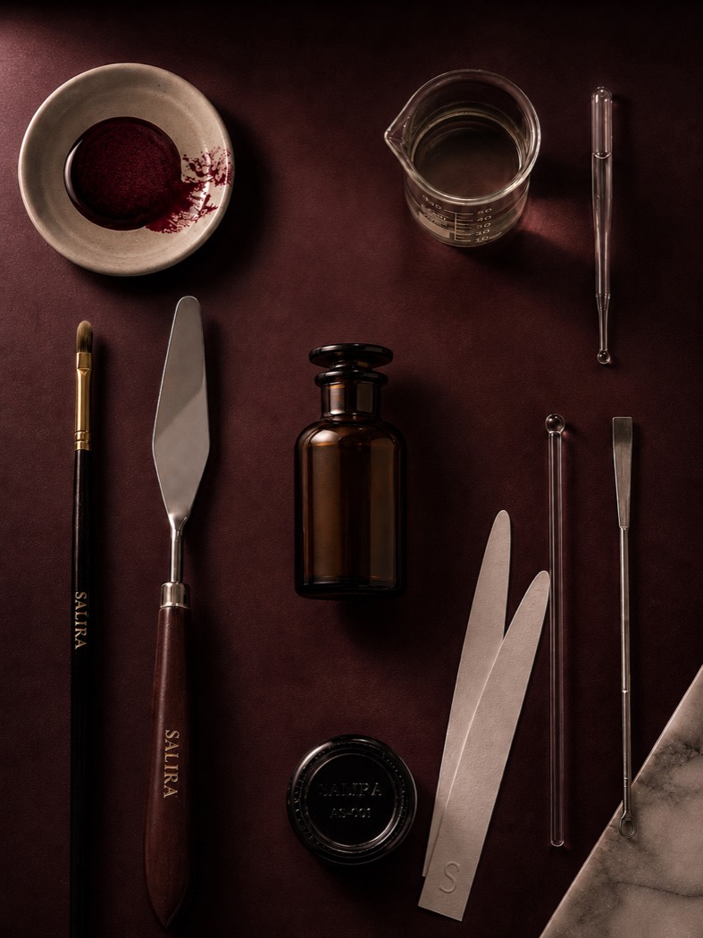A flat-lay of Salira's perfumer's tools on a deep burgundy surface — a Salira-engraved brush and palette knife, an amber apothecary bottle, a glass beaker and pipette, and metal blending spatulas