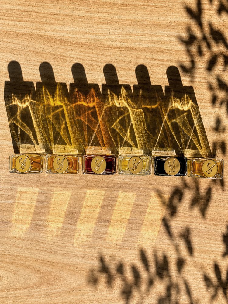 Six rectangular Salira fragrance bottles with engraved gold medallion caps arranged in a row on pale wood, casting long shadows in dappled light