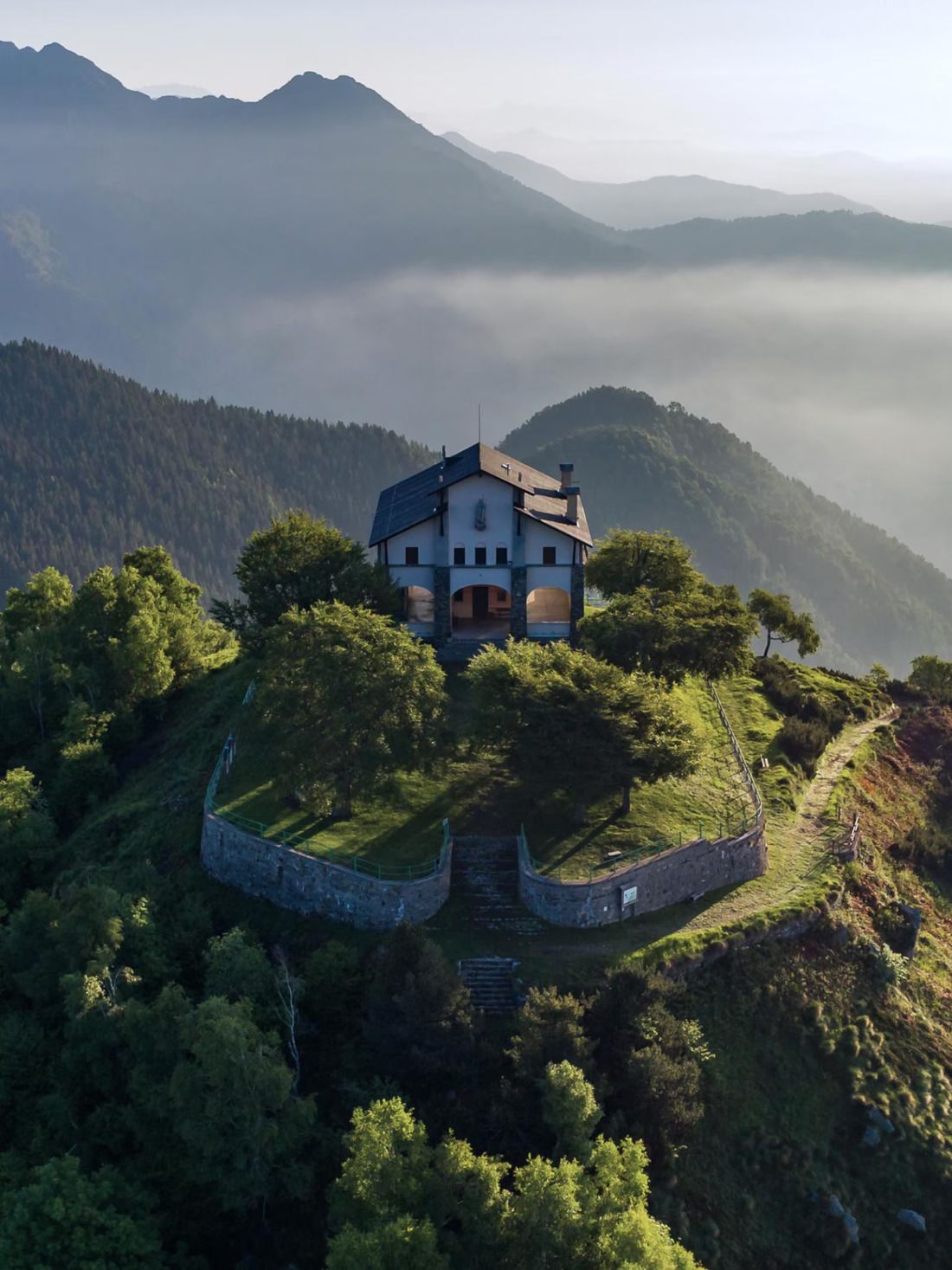 A drone shot of a stunning family home atop a hill surrounded by greenery, with mountain vistas in the background